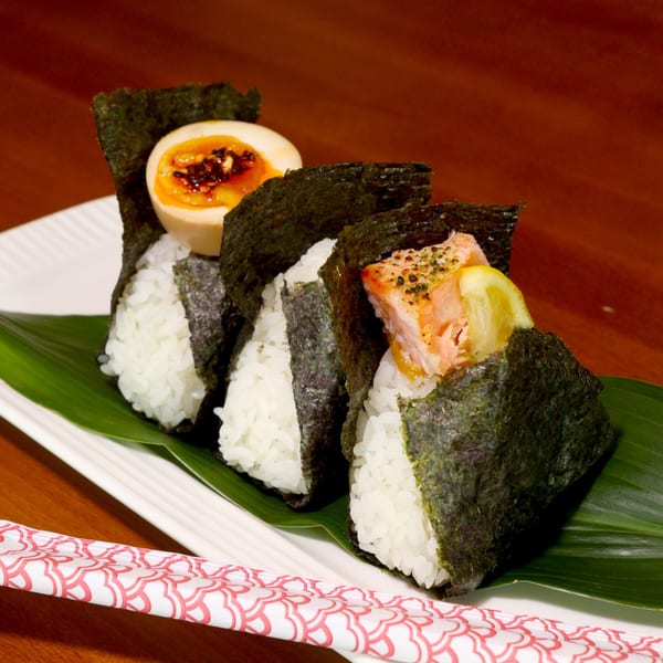Three Tiger Ramen onigiri, one with a soft-boiled egg, another with salmon and lemon, on a white plate.
