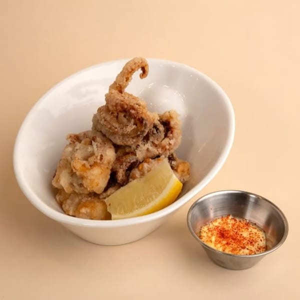 Crispy fried calamari with a lemon wedge in a white bowl, alongside a metal cup of dipping sauce.