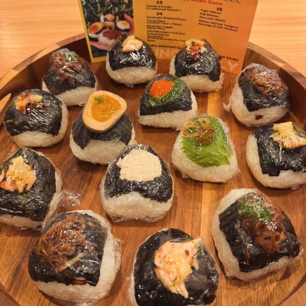 A wooden tray holds a variety of Tiger Ramen onigiri, individually wrapped in plastic, with a menu in the background.