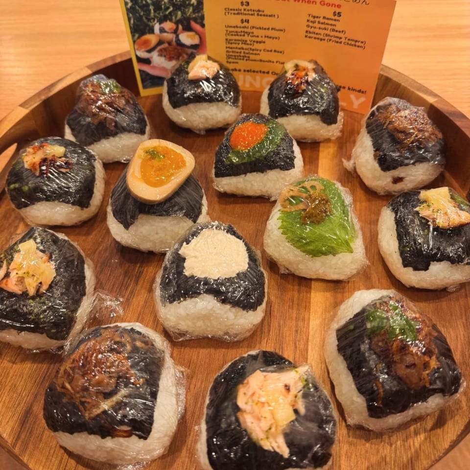 A wooden tray holds a variety of Tiger Ramen onigiri, individually wrapped in plastic, with a menu in the background.