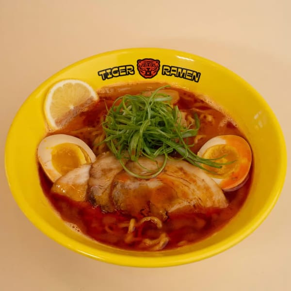 A vibrant yellow bowl of Tiger Ramen with pork, soft-boiled eggs, green onions, and a lemon slice.