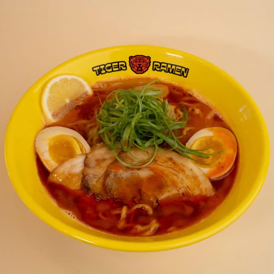 A vibrant yellow bowl of Tiger Ramen with pork, soft-boiled eggs, green onions, and a lemon slice.