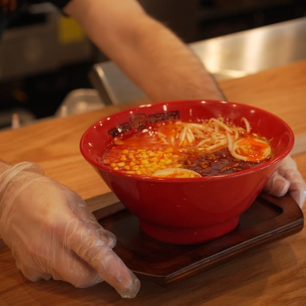 A server wearing gloves places a red bowl of Tiger Ramen with corn, sprouts, and egg on a wooden tray.