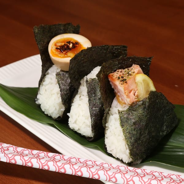 Three onigiri with salmon, egg, and plain rice, wrapped in nori, on a white plate with chopsticks.